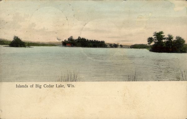 Hand-colored view across water towards three islands in big Cedar Lake. The center island has a resort building with a red roof.