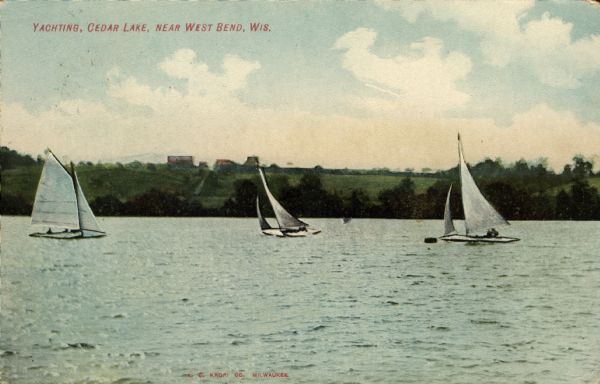 Hand-colored view across water of three, two-person sailboats on Cedar Lake. On the far shoreline are buildings at the top of a hill. Caption reads: "Yachting, Cedar Lake, near West Bend, Wis."