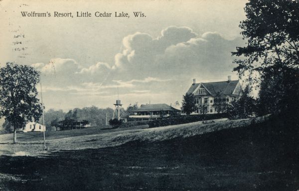 View up sloped lawn towards Wolfrum's Resort at Cedar Lake. Along the top of the slope are four buildings and a water tower. Caption reads: "Wolfrum's Resort, Little Cedar Lake, Wis."
