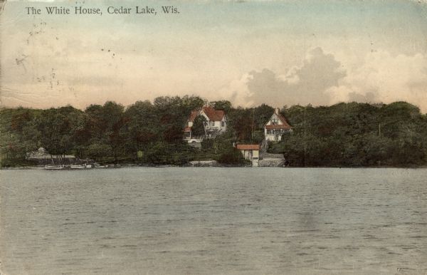 Hand-colored view of the White House resort at Cedar Lake. The view is across water towards two buildings on a wooded hill, and a dock with boats, and a boat shelter. Caption reads: "The White House, Cedar Lake, Wis."