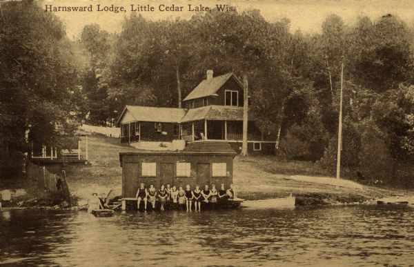 View across lake towards Harnsward Lodge, with a group of people in bathing suits sitting on the landing of the boathouse. Caption reads: "Harnsward Lodge, Little Cedar Lake, Wis."