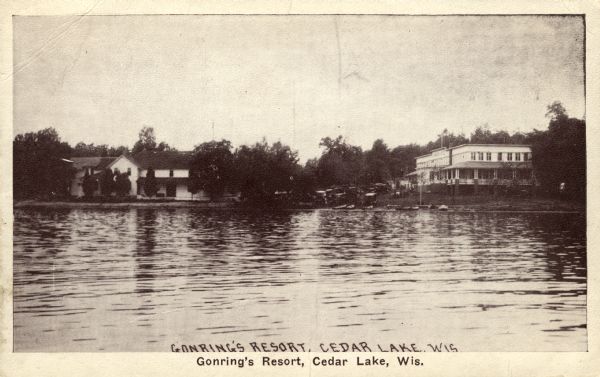 View across lake towards the Gonring's Resort from Cedar Lake. Along the shoreline are two buildings, a parking lot and a boat landing. Caption reads: "Gonring's Resort, Cedar Lake, Wis."