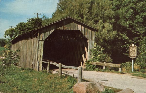 View from side of road towards the covered bridge, with the historical marker on the right. "This bridge was built by the town of Cedarburg on petition of neighboring farmers to replace periodically washed out bridges. Built in 1876, retired in 1962. The Ozaukee County Board assumes the preservation and maintenance."