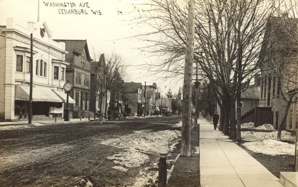Photographic postcard view down unpaved Washington Avenue. A clock on the sidewalk on the left is in front of a musical instrument shop. Snow is on the ground, and pedestrians are on the sidewalks and in front of shops. Caption reads: "Washington Ave., Cedarburg, Wis."