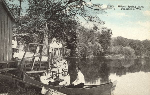 Hand-colored view along shoreline of people posing in a rowboat near a boathouse. Other people are leaning against a railing and standing near a boathouse. Caption reads: "Hilgen Spring Park, Cedarburg, Wis."
