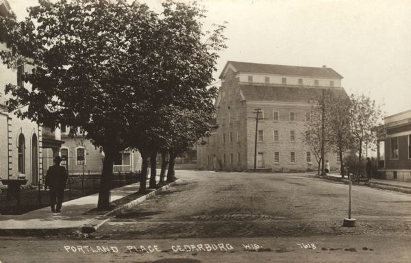 Photographic postcard view across intersection towards Portland Place, a five-story stone building. There are pedestrians on the sidewalks on the left and right in the foreground. Caption reads: "Portland Place, Cedarburg, Wis."