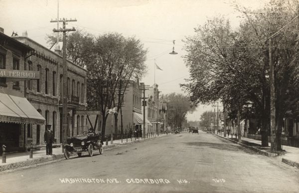 Photographic postcard of Washington Avenue featuring the Union House on the left, as well as a furniture store and garage. Automobiles are parked in the street. A street lamp is hanging from the power line. Otto Beckmann, Chief of Police, is identified as the man on the sidewalk. Caption reads: "Washington Ave., Cedarburg, Wis."