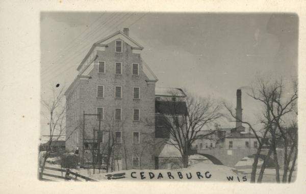 Photographic postcard of a mill next to Cedar Creek. There is a bridge on the right, and a factory in the background..