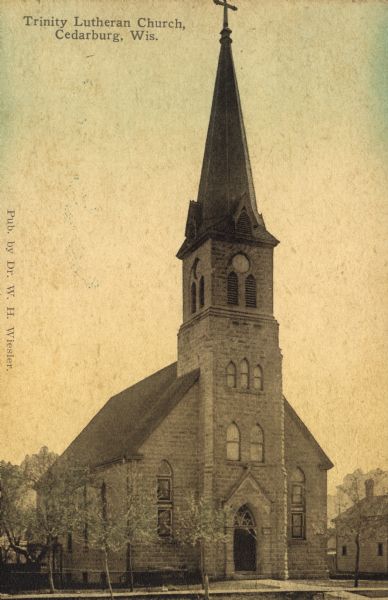 Hand-colored view of Trinity Lutheran Church on Columbia Road, a stone building with a clock in the bell tower/steeple. Arched windows are on the front and sides of the church. Caption reads: "Trinity Lutheran Church, Cedarburg, Wis."