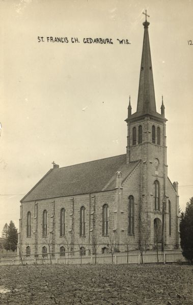 View toward the church, which is a stone building with a clock in the bell tower/steeple. Arched windows are on the front and sides. A fence is running along the yard. Caption reads: "St. Francis Ch., Cedarburg, Wis."