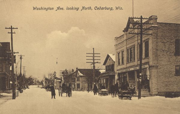 View of Washington Avenue on a snowy day. The A.F. Bruss Building is on the right. Horse-drawn carts and sleighs are in the street. Caption reads: "Washington Ave. looking North, Cedarburg, Wis."