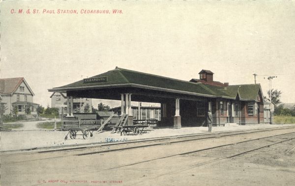 Hand-colored view of the train station. Two carts and a man are standing on the platform. Railroad tracks are in the foreground, and houses are in the background. Caption reads: "C. M. and St. Paul Station, Cedarburg, Wis."