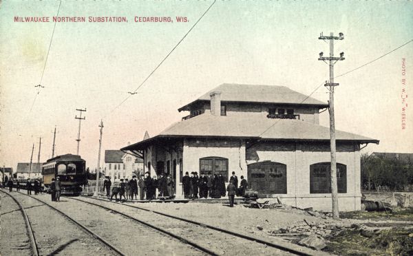 Hand-colored view down railroad tracks toward the substation. An interurban car is on the railroad tracks, and a group of passengers are on the platform. Caption reads: "Milwaukee Northern Substation, Cedarburg, Wis."