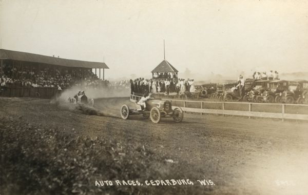 View of an auto race with cars on the track, a crowd in the grandstand, and other spectators and their cars in the center oval. Caption reads: "Auto Races, Cedarburg, Wis."