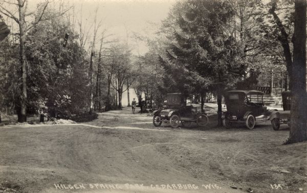 Photographic postcard view of two men standing on a tree-lined road leading to water. Automobiles are parked on the right near a gazebo. Caption reads: "Hilgen Spring Park, Cedarburg, Wis."