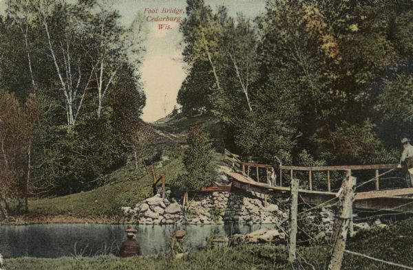 Hand-colored view of a footbridge crossing over Cedar Creek. A man is fishing from the bridge, and a dog is standing nearby. Two boys are sitting in the grass.