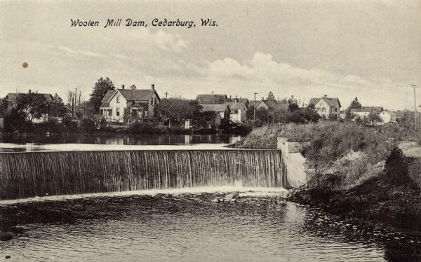 Photographic postcard view of the Woolen Mill Dam. Several dwellings are in the distance. Caption reads: "Woolen Mill Dam, Cedarburg, Wis."