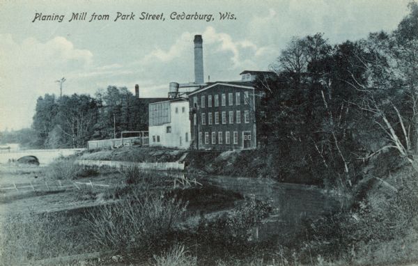 Tinted view of the Planing Mill, with Cedar Creek running alongside. There is a bridge on the left. Caption reads: "Planing Mill from Park Street, Cedarburg, Wis."