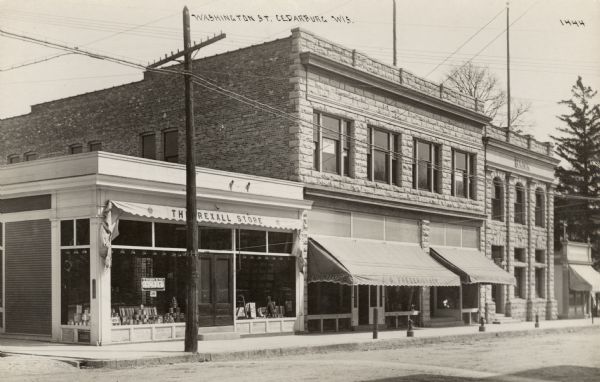 View from street of the Washington Street block with, from left, the Rexall Store, J.G. Fredericksen Furniture, and a bank. A rack of postcards is on display in the Rexall window. Hitching posts are along the sidewalk. Caption reads: "Washington St., Cedarburg, Wis."