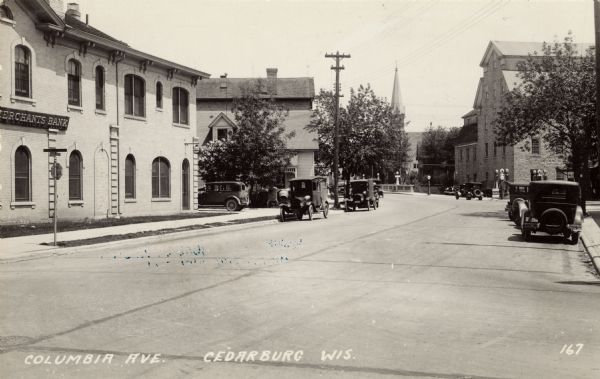 View looking up Columbia Avenue, with the Merchants Bank on the left, and a mill, and a church further down on the right. Automobiles are parked along the curbs. There are Mobiloil gas pumps next to the mill. Caption reads: "Columbia Ave., Cedarburg, Wis."