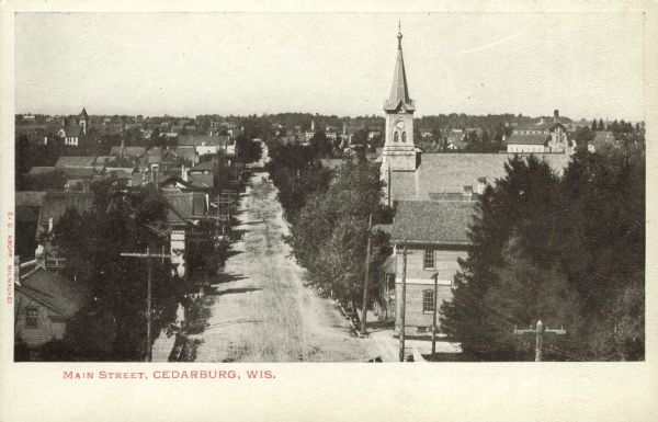 Elevated view looking down Main Street. A church is on the right, surrounded by dwellings obscured by trees. A mill is in the distance. Caption reads: "Main Street, Cedarburg, Wis."