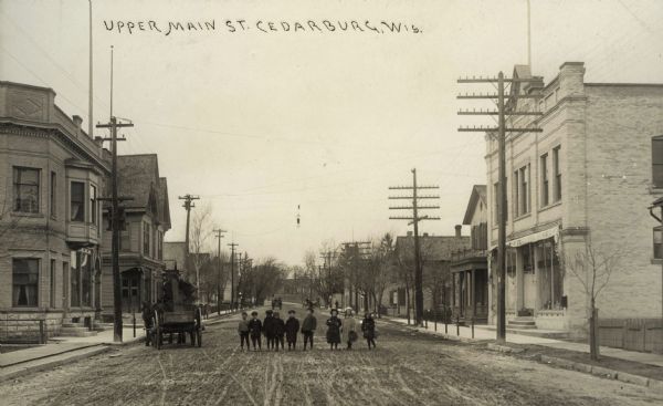 Photographic postcard view looking down the center of Main Street towards a group of children standing in a row in the center of the street. There is a horse-drawn cart near the group on the left. Caption reads: "Upper Main St., Cedarburg, Wis."