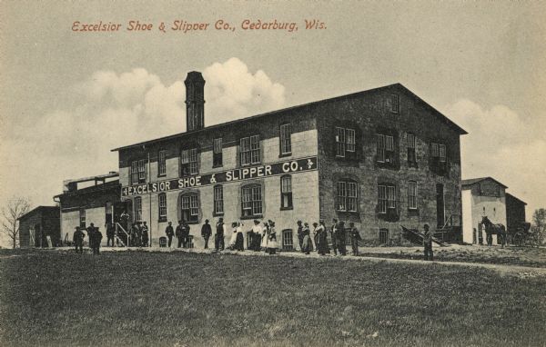 Exterior view across lawn toward a group of people standing on the front walkway of the building. A horse and buggy are parked near the back of the building on the right. Caption reads: "Excelsior Shoe & Slipper Co., Cedarburg, Wis."