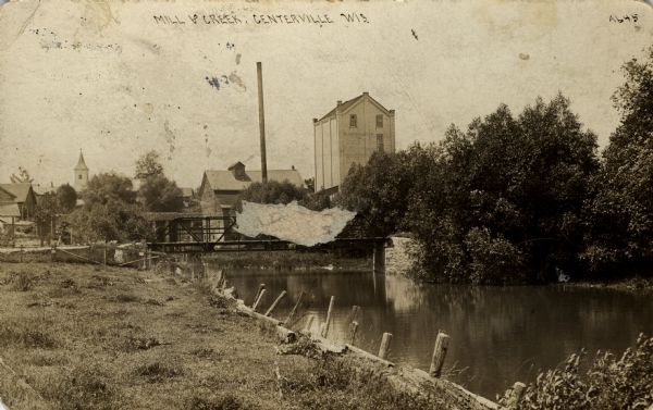 Photographic postcard view of the mill and creek, with a bridge in front of the mill. The skyline of Centerville is in the background. Caption reads: "Mill and Creek, Centerville, Wis."