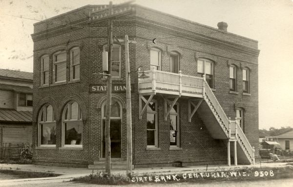 Exterior view from street of the State Bank with a corner entrance. The brick building has two-stories, with a set of wooden stairs on one side leading up to the second floor. Caption reads: "State Bank, Centuria, Wis."
