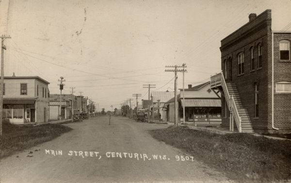 View down center of Main Street. The State Bank is in the foreground on the right. A hardware store and hotel are on the left. There is a windmill in the distance. Caption reads: "Main Street, Centuria, Wis."
