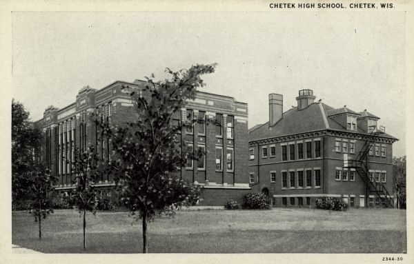 View across lawn towards Chetek High School. Two buildings are on the grounds. The one on the right has a fire escape and gabled roof. Caption reads: "Chetek High School, Chetek, Wis."