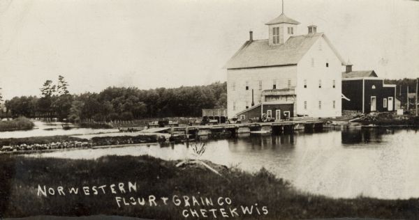 View from shoreline across water towards the Norwestern Flour and Grain Company mill on the banks of a river. Caption reads: "Norwestern Flour and Grain Co. Chetek, Wis."
