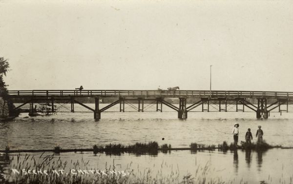 View from shore towards a bridge crossing a river. Three boys are standing in the marshy foreground, and one of them has a fishing pole. A horse and buggy are crossing the bridge, and a man on the bridge is lifting his leg above the railing. Caption reads: "A Scene at Chetek, Wis."