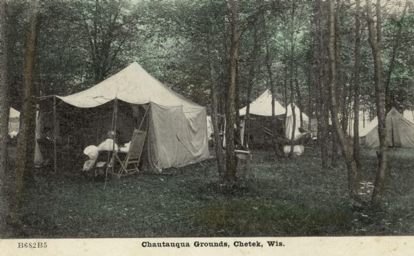 Hand-colored scene of a campsite in the woods with several tents. Men are sitting in chairs in the tent on the left. Caption reads: "Chautauqua Grounds, Chetek, Wis."