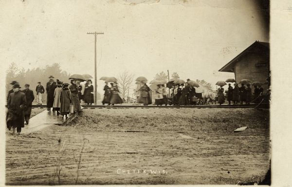 View across road towards the Chetek train depot on a rainy day. People are walking along the platform and walkway, some carrying umbrellas. Caption reads: "Chetek, Wis."