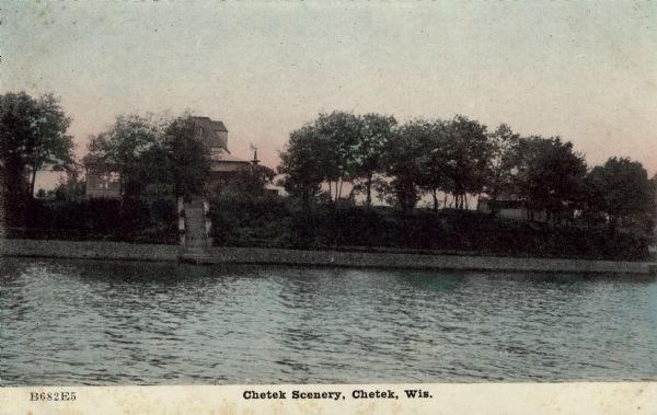 Hand-colored postcard view across water towards the shoreline of Lake Chetek. Along the shoreline is a stone retaining wall and a flight of steps leading from the lake up to a house and other buildings. Caption reads: "Chetek Scenery, Chetek, Wis."