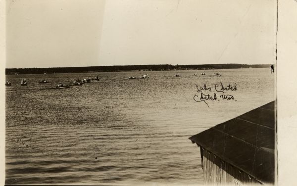 Elevated view from shoreline looking over the roof of a building in the right foreground towards people in rowboats on Lake Chetek. The far shoreline is the the distance. Caption reads: "Lake Chetek, Chetek, Wis."