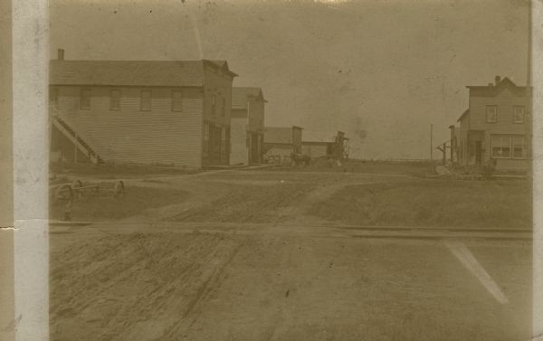 View down unpaved street in Chili towards commercial buildings, with horses and wagons parked on the street. Railroad tracks are in the foreground.