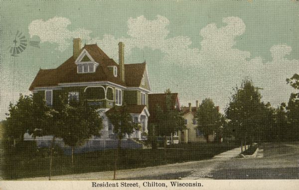 Hand-colored photomechanical view of a residential neighborhood with a large house on the corner. Trees line the street, and there is a windmill on the left. Caption reads: "Resident Street, Chilton, Wisconsin."