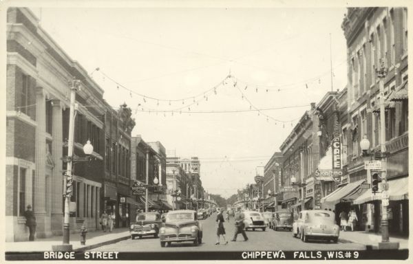 Looking north on Bridge Street from Spring Street intersection. In foreground, left, is a bank. In foreground, right, is a restaurant with a neon sign that reads: "Skogmo Cafe". The view includes pedestrians on the sidewalks and crossing the street, parked cars and moving cars, and canvas awnings on many stores covering sidewalks. Caption reads: "Bridge Street, Chippewa Falls, Wis."