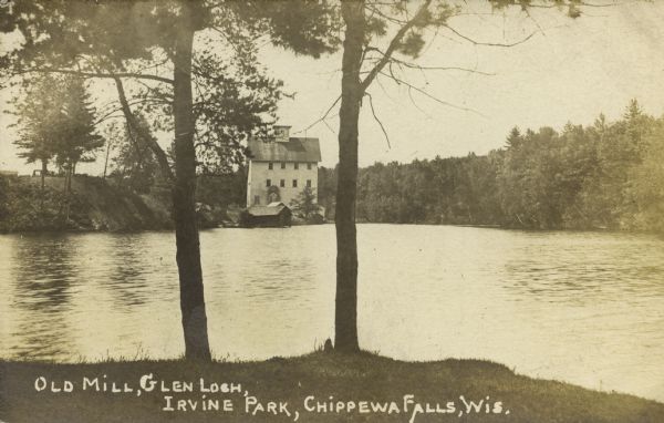 View from opposite shoreline through trees toward the Glen Loch dam and mill in Irvine Park. Caption reads: "Old Mill, Glen Loch, Irvine Park, Chippewa Falls, Wis." 