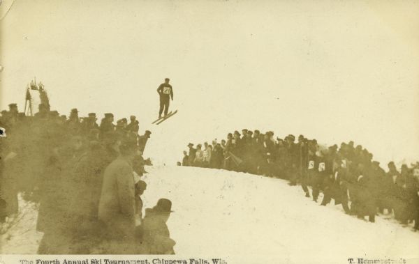 View looking up towards a ski jumper airborne over the top edge of a hill as a large crowd is looking on from the sides. The top of the ski jump scaffold is in the far background. Caption reads: "The Fourth Annual Ski Tournament, Chippewa Falls, Wis."