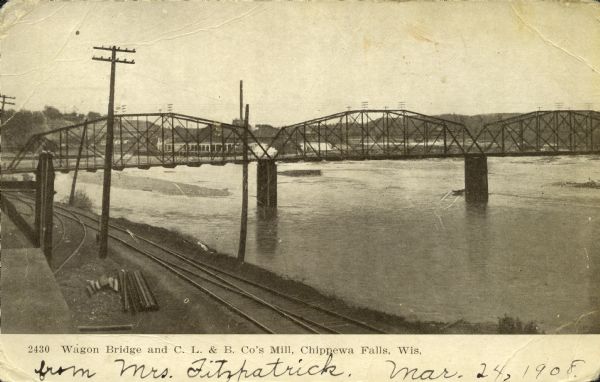 Elevated view looking east and upstream from the shoreline towards the wide Chippewa River. The water level is nearly up to the railroad tracks in the foreground on the north shore. The Big Mill is visible through the steel beams of the Wagon Bridge. Caption reads: "Wagon Bridge and C. L. & B. Co's Mill, Chippewa Falls, Wis."