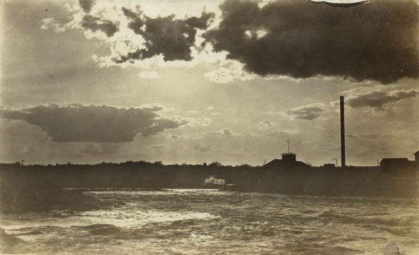 View from the south shore of the Chippewa River, looking west, showing choppy, churning rapids, and a silhouette of the Big Mill and smokestack.