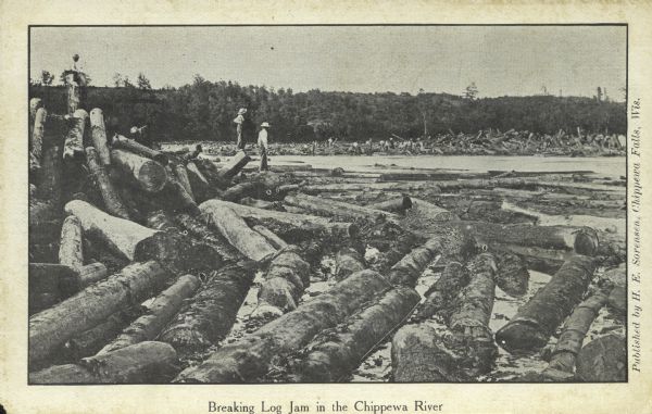 Two men standing on logs are looking at the log jam in the river. Caption reads: "Breaking Log Jam in the Chippewa River"