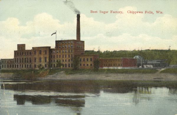 View across the Chippewa River towards an industrial red brick building, that is six stories tall in the center section, with a large smokestack at the center. Caption reads: "Beet Sugar Factory, Chippewa Falls, Wis."