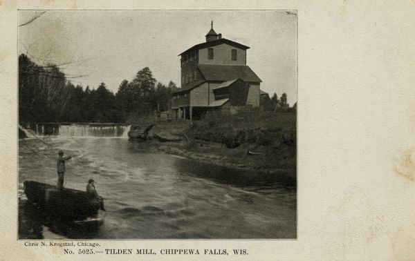 View of Tilden Mill and dam on Duncan Creek. In the foreground two men are standing and sitting on a large, flat boulder. Caption reads: "Tilden Mill, Chippewa Falls, Wis."