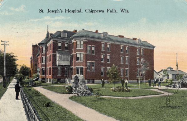 Hand-colored postcard view of St. Joseph's Hospital. The view is facing south-west and reveals a large fenced side lawn with foot paths, plants and six people. On the right are two service buildings. On the left, across a street are the two steeples of St. Charles Catholic church. Caption reads: "St. Joseph's Hospital, Chippewa Falls, Wis."