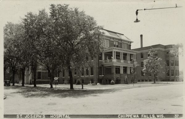 View from street towards the St. Joseph's Hospital. In the center is a two level open porch, with two people on the second level. A 1930s automobile is parked by the front door. A road sign reads: "Quiet Hospital Zone."