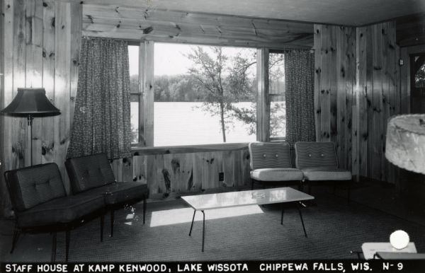 Interior view of a living room in the Staff House at Kamp Kenwood, Lake Wissota. There is a view of Lake Wissota through the large window. Caption reads: "Staff House at Kamp Kenwood on Lake Wissota, Chippewa Falls, Wis."